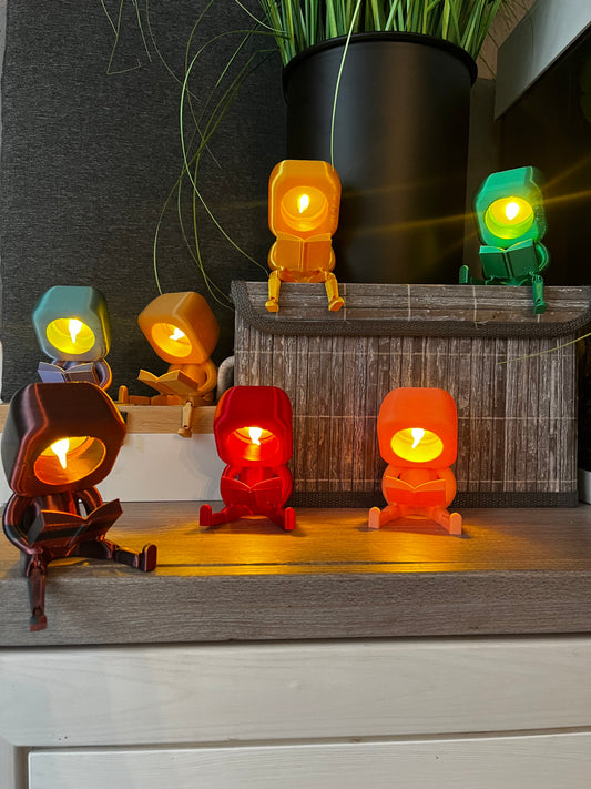 Mehrere 3D-gedruckte lesende Gefährten als Nachtlicht in verschiedenen Farben, dekorativ arrangiert auf einem Regal, aus PLA, perfekte Geschenkidee für Buchliebhaber und Kinderzimmer, stimmungsvolle Beleuchtung, Club der lesenden Gefährten