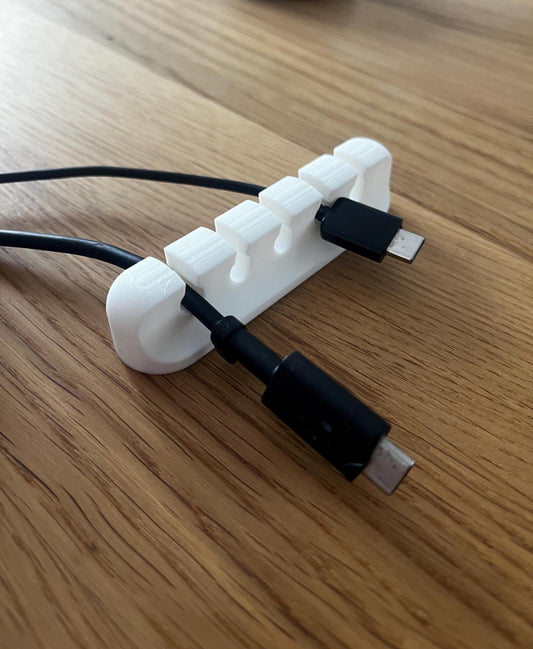 Weißer 3D‑gedruckter Kabelorganisierer mit fünf Schlitzen liegt auf einer Holzoberfläche; zwei schwarze USB‑C‑Ladekabel sind in den vorderen Öffnungen eingeclipst und laufen nach links aus dem Bild. Abgerundete Kanten, kompakte Bauform, Fokus auf ordentliche Kabelführung am Schreibtisch oder Sideboard.