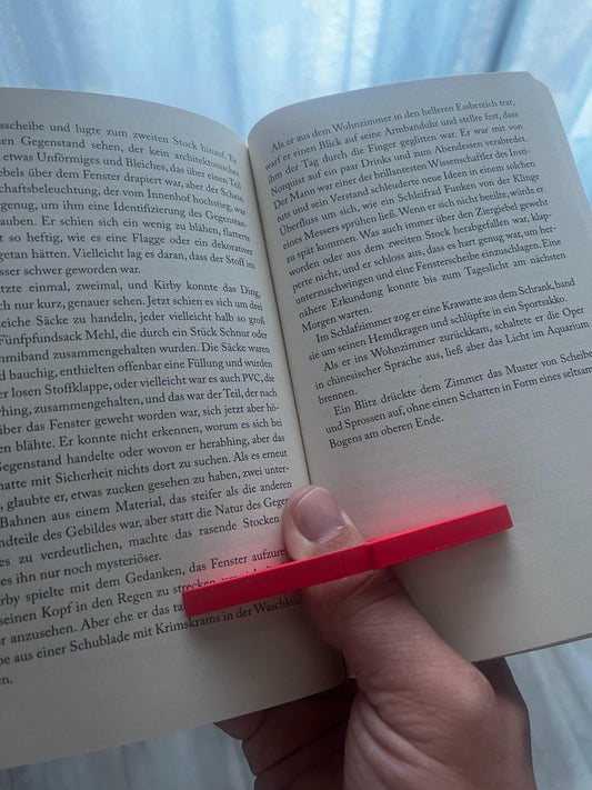 Hand hält offenes Buch mit rotem 3D‑gedruckten Buchhalter am Daumen; Seiten bleiben für einhändiges Lesen offen.