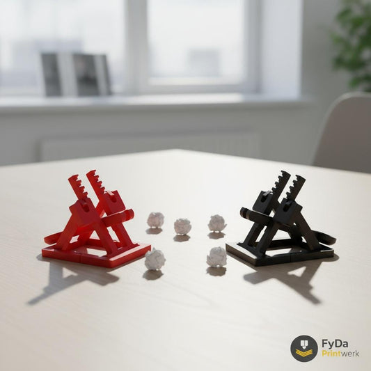 Zwei Katapulte (Rot & Schwarz) auf Schreibtisch, Papierkügelchen (Mockup):
Zwei 3D-gedruckte Katapulte in Rot und Schwarz, gegenüberstehend auf Schreibtisch mit Papierkügelchen, FyDa Printwerk